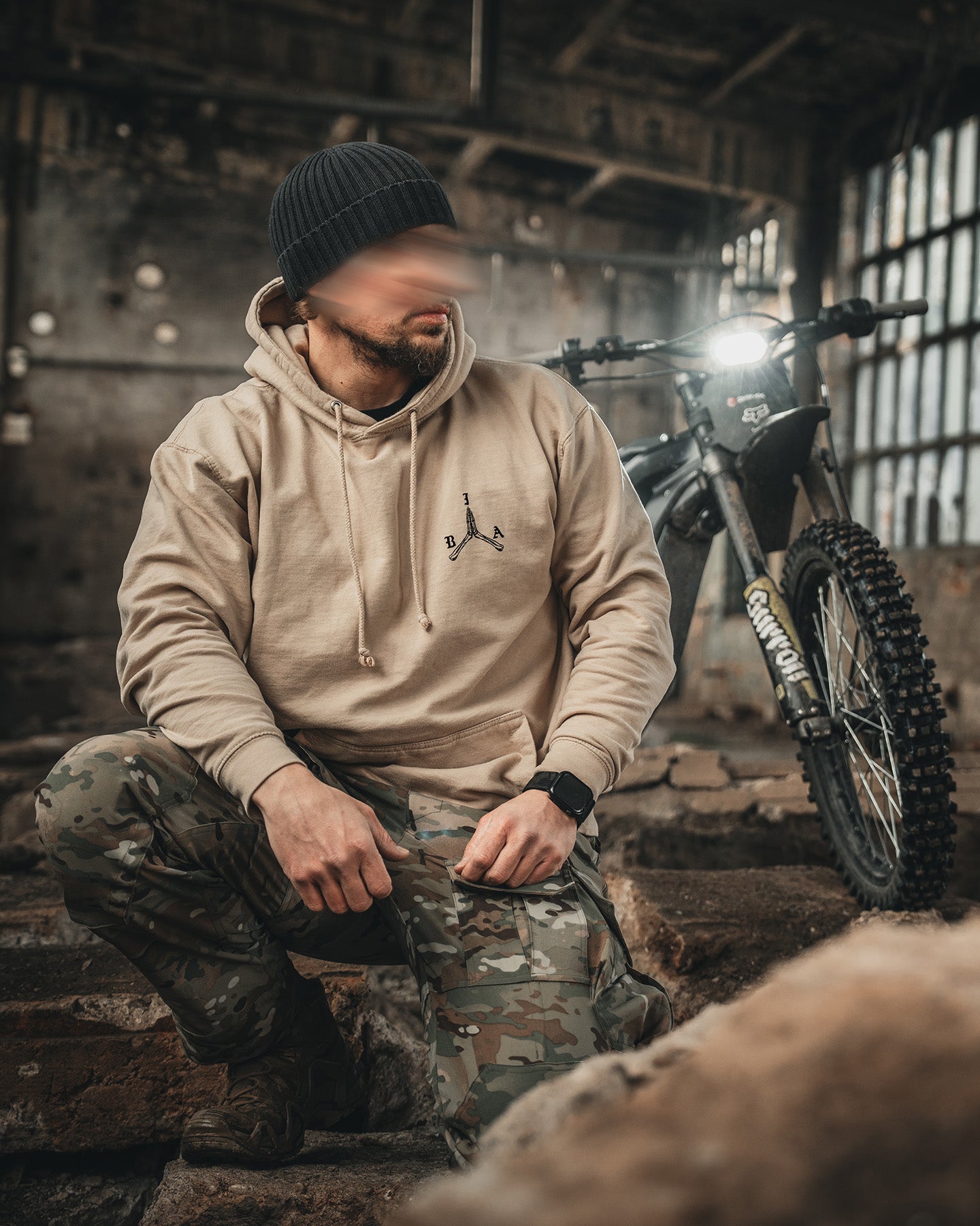 Man crouching beside a motorcycle in the sand Stay Violent Hoodie #color_sand #in_field