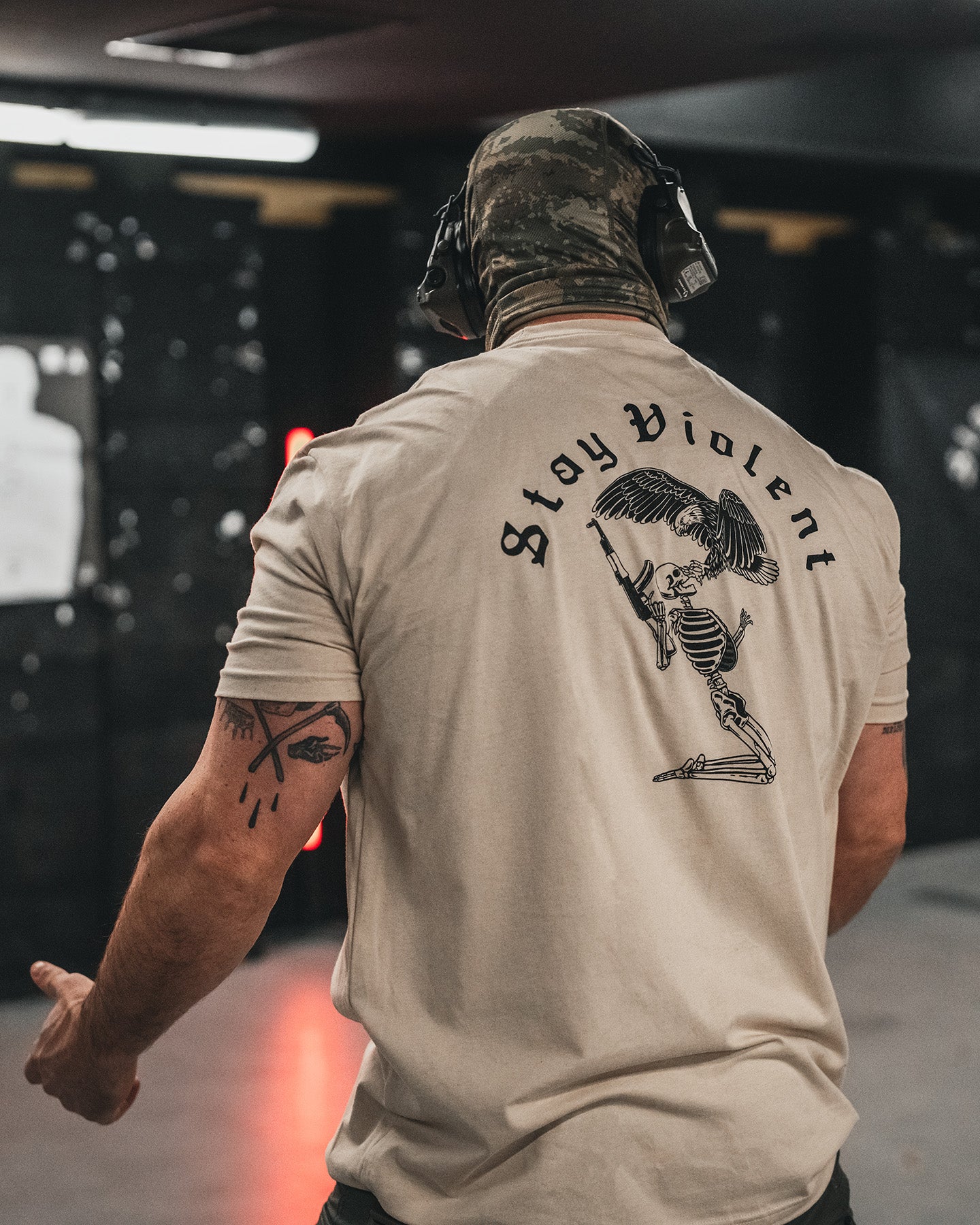 Man from behind in the sand Stay Violent T-Shirt at an indoor shooting range, wearing ear protection #color_sand #in_field