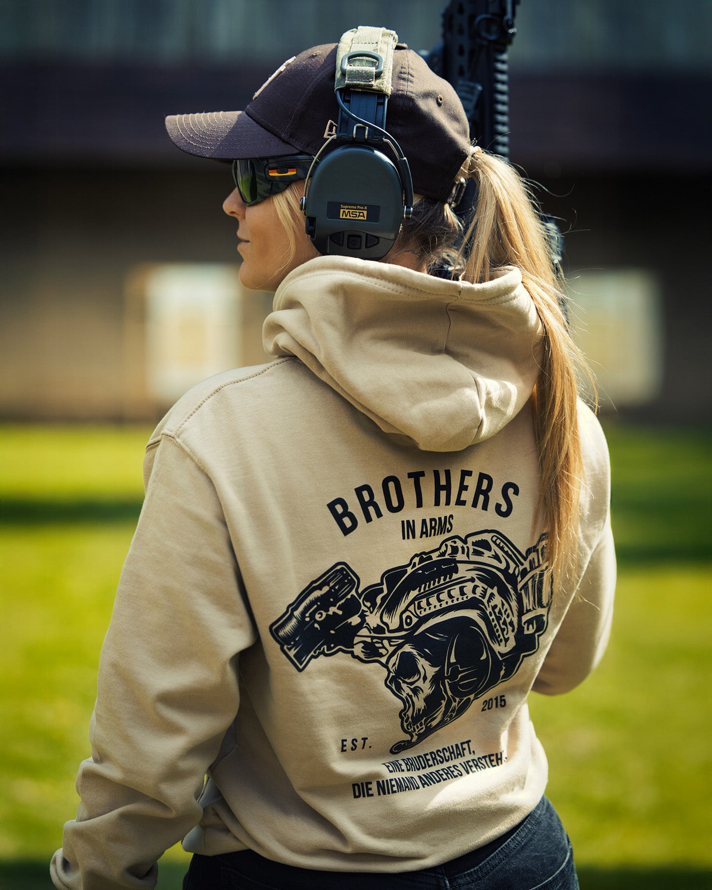Woman from behind in the sand Team Hoodie at a shooting range, showing the back print #color_sand #in_field