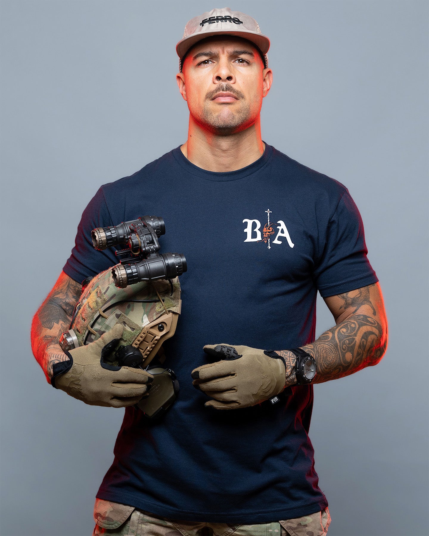 Man wearing a navy blue t-shirt with a logo, holding tactical gear against a gray background. #color_navy
