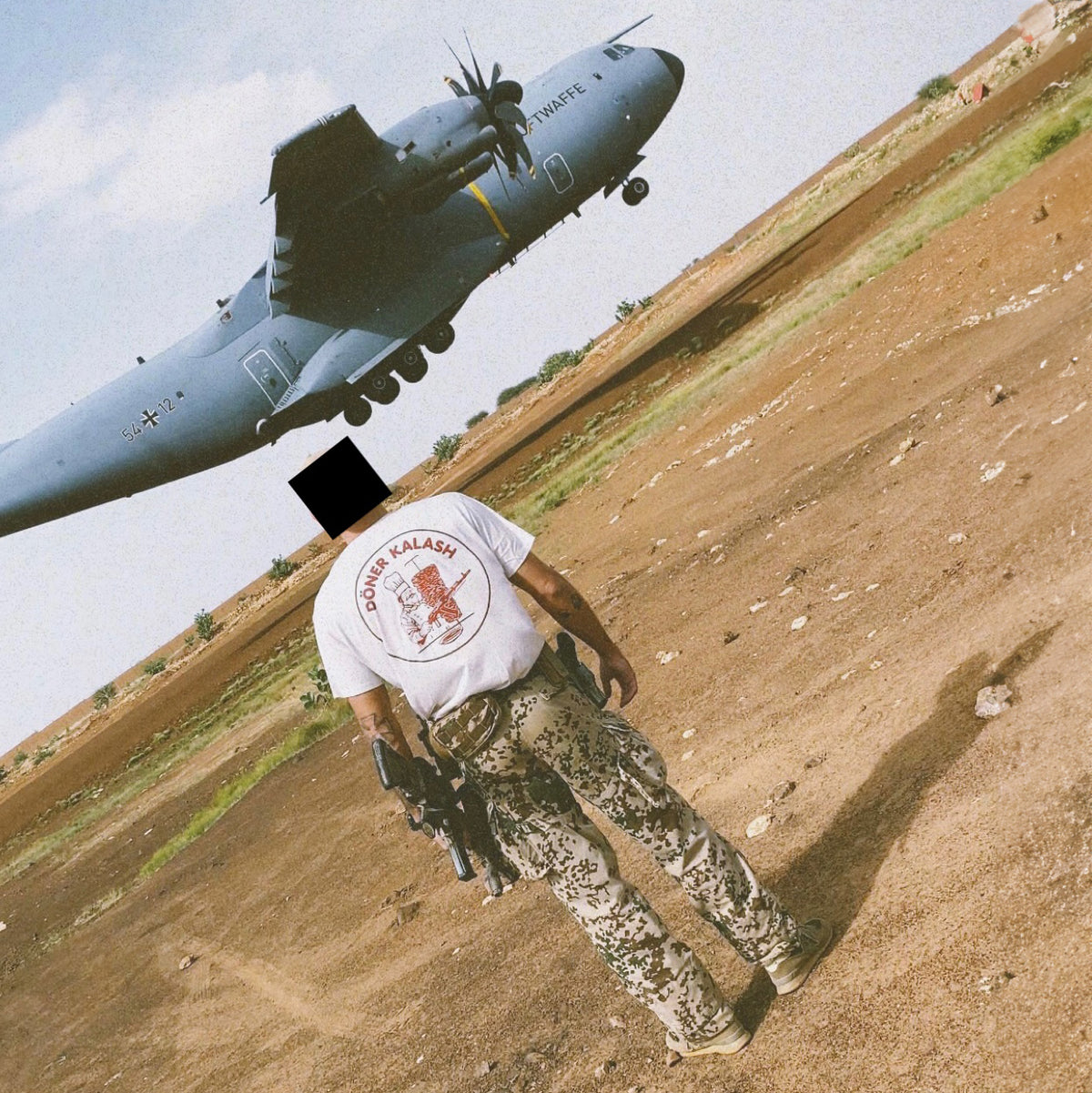 Man from behind in a white Döner Kalash shirt on sand, military transport aircraft behind him