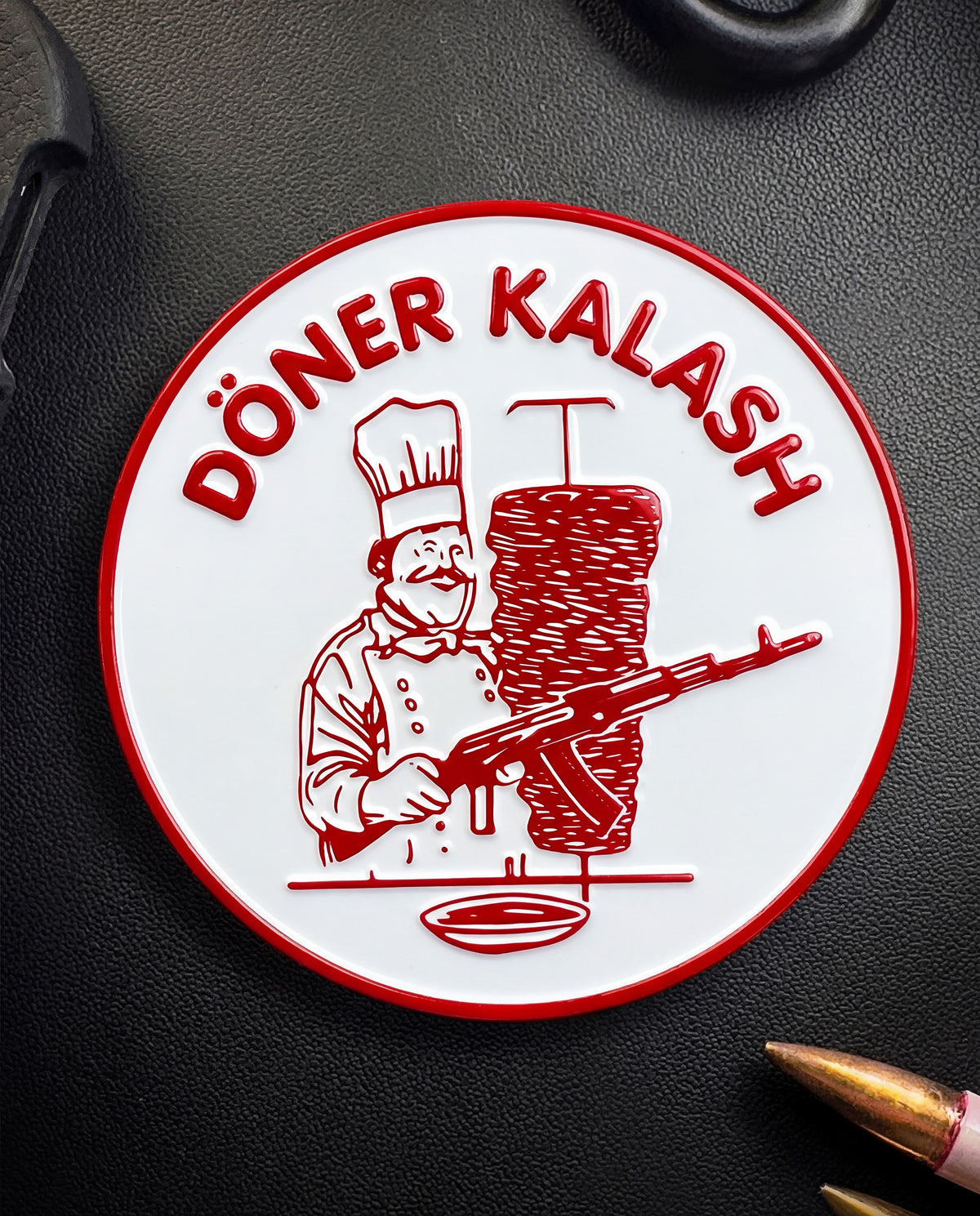 Round Döner Kalash coin in red and white on a dark surface next to a rifle cartridge