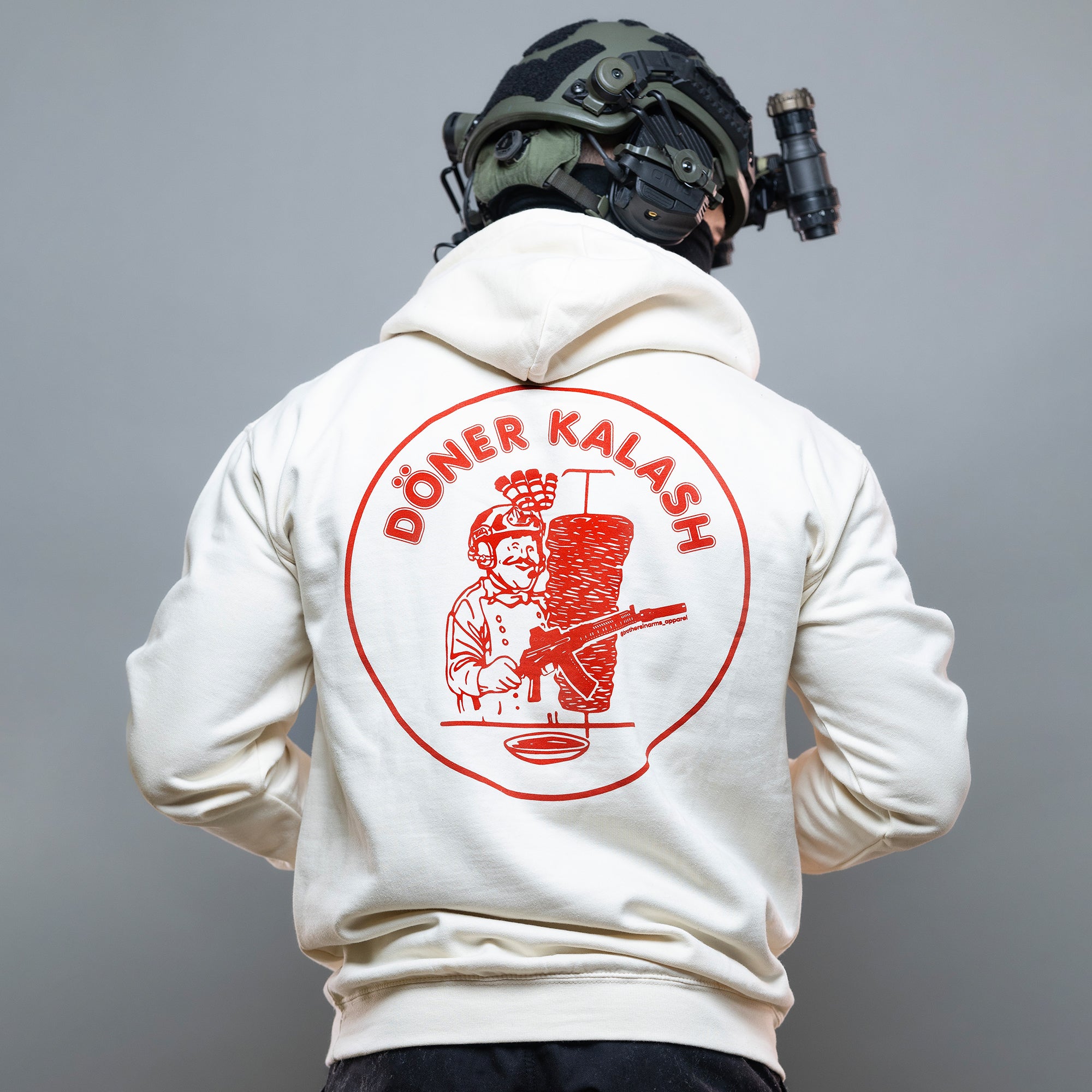 Eine Person trägt einen taktischen Helm und den „Döner Kalash Spicy Hoodie“ von Brothers in Arms mit einer farbenfrohen Döner-Illustration auf der Rückseite.