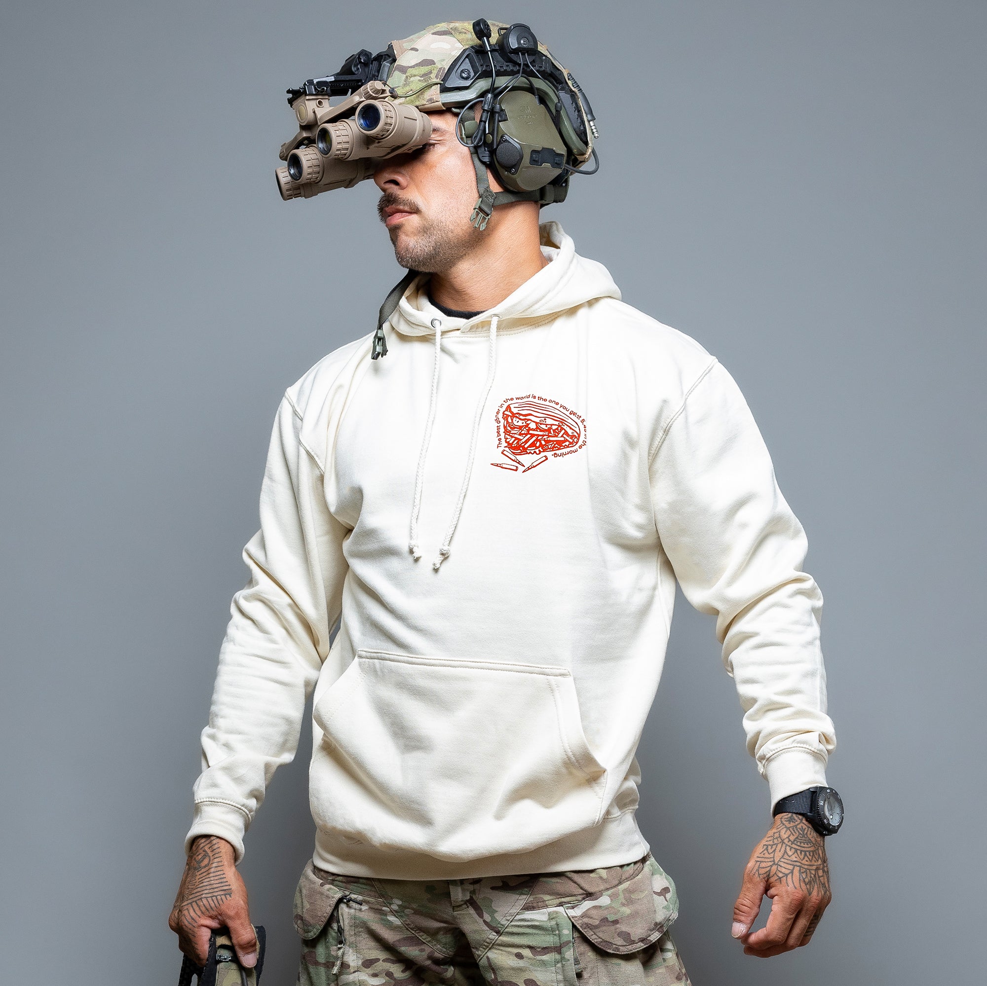 Die Person trägt einen Döner-Kalash-Kapuzenpullover von Brothers in Arms und Tarnhosen sowie taktische Ausrüstung wie Schutzbrillen und Helme und präsentiert damit die urbanen Stiltrends Deutschlands vor einem grauen Hintergrund. #color_cream