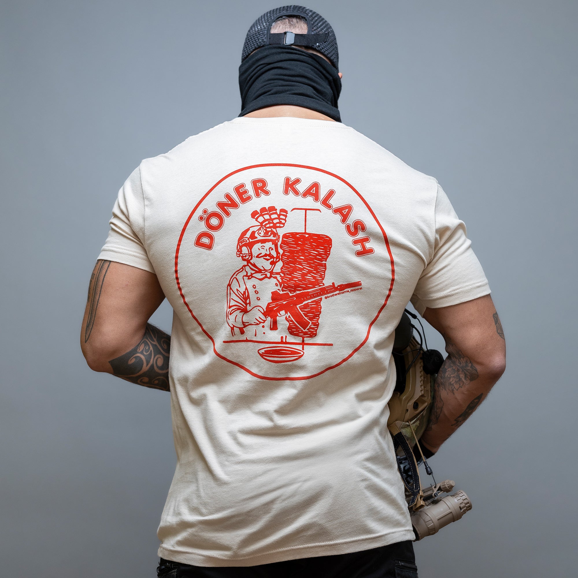 Eine Person trägt ein „Döner Kalash Spicy T-Shirt“ von Brothers in Arms, auf dem ein Koch mit einem Gewehr abgebildet ist, und das den kantigen Vibe der Spicy Edition verkörpert. #color_sand