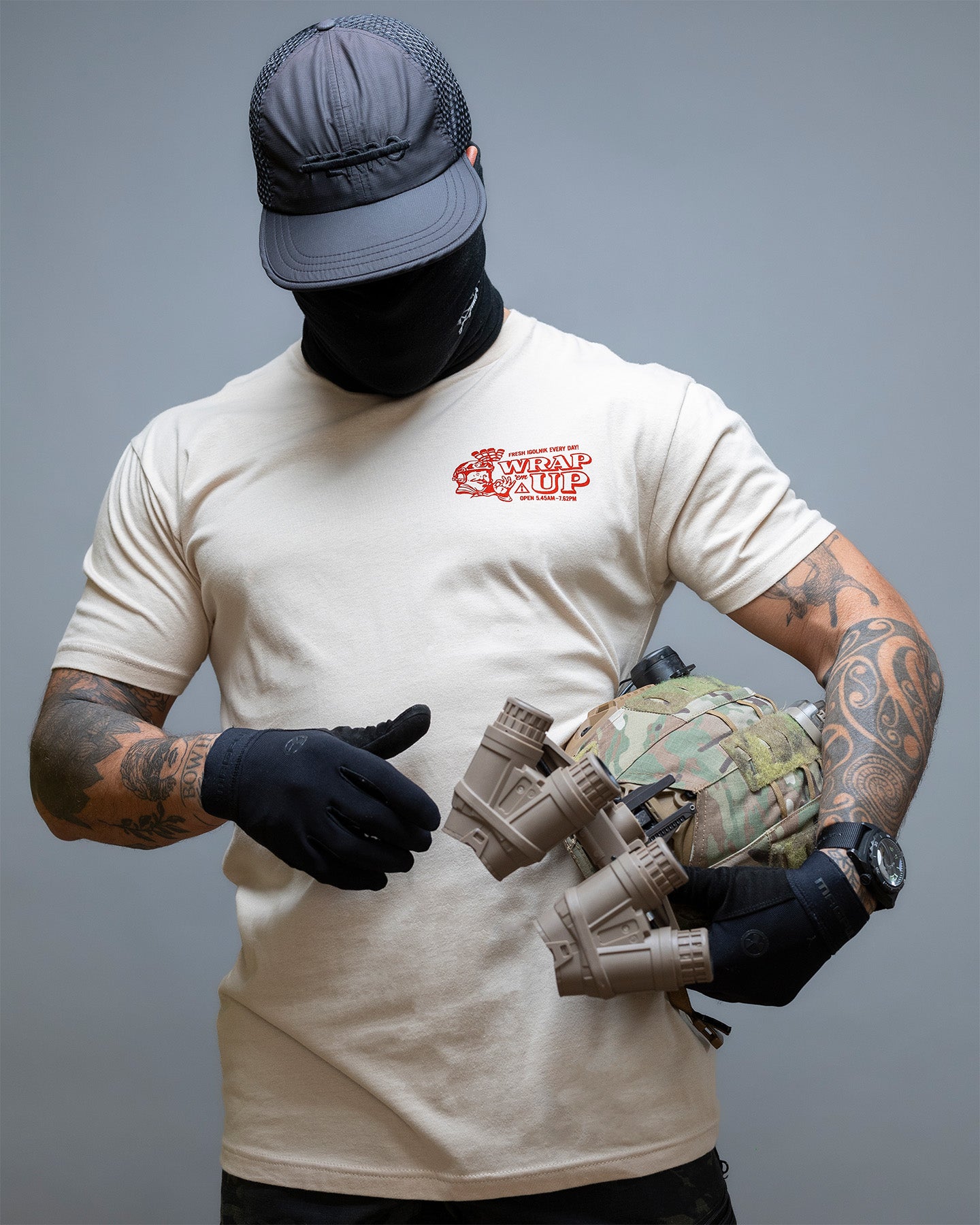 Person wearing a tan t-shirt with a logo, holding camouflage gear against a gray background. #color_sand
