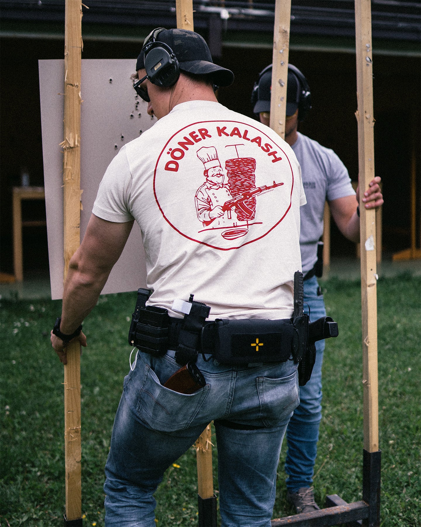 Person wearing a 'Döner Kalash' t-shirt with a graphic design, holding wooden sticks outdoors. #color_white #in_field