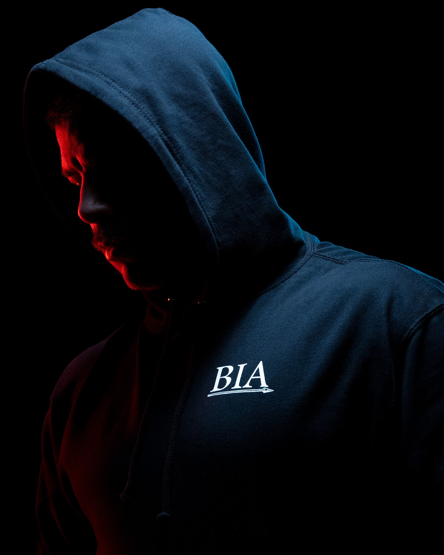 Man in the black Comfort Is A False God Hoodie with the hood up, lit red from behind #color_black #in_field