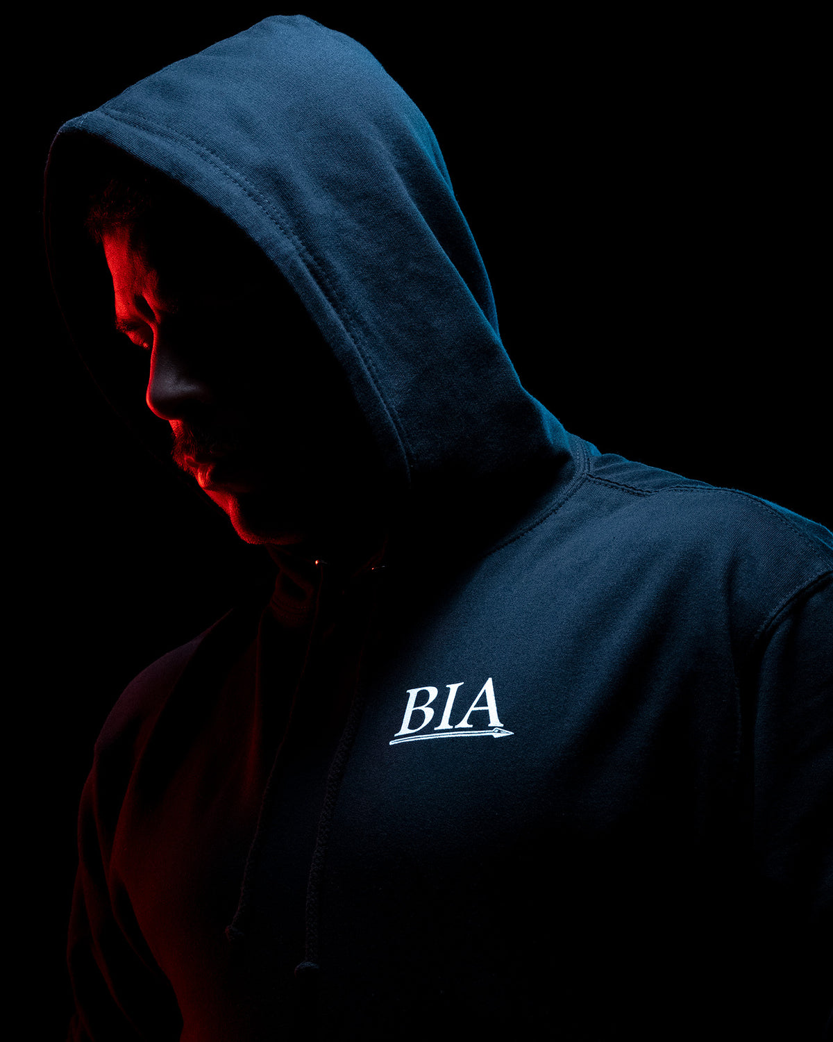 Man in the black Comfort Is A False God Hoodie with the hood up, lit red from behind