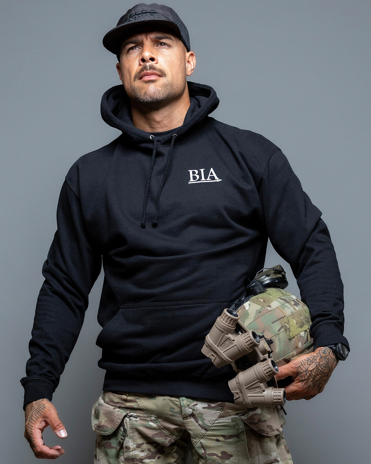 Man in a cap wearing the black Comfort Is A False God Hoodie, front view with the BIA chest print