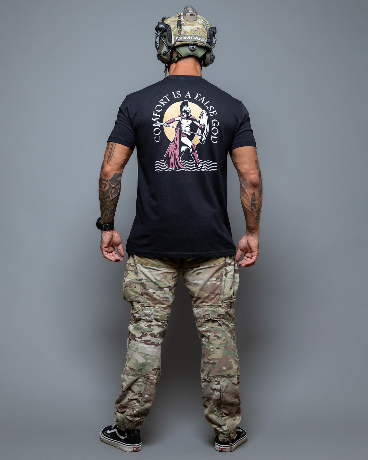 Full-length shot of the black Comfort Is A False God T-Shirt from behind, worn with camo trousers