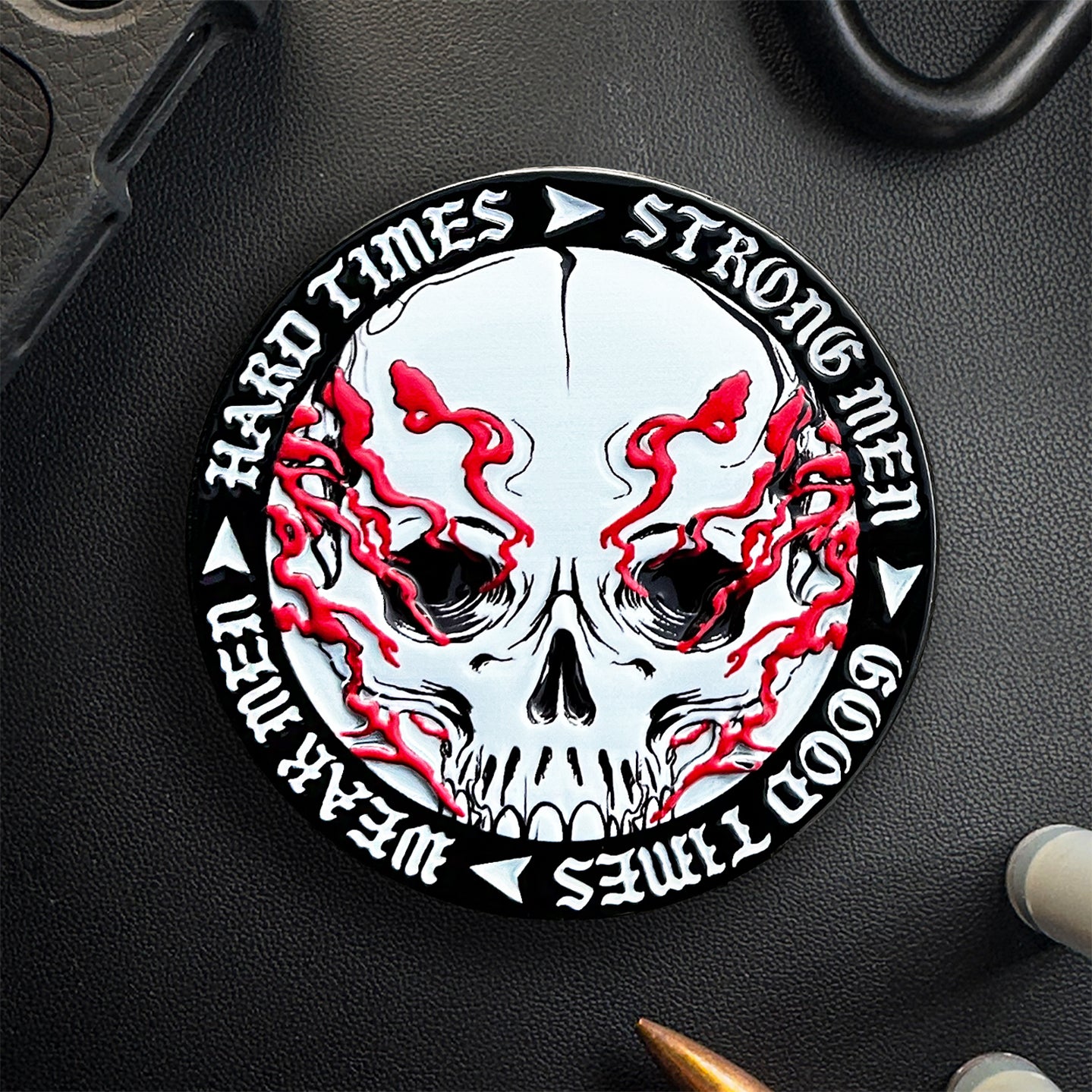 Die „Hard Times Coin“ von Brothers in Arms ist ein runder Abzeichen mit einem von roten Flammen umhüllten Totenkopf, umrandet vom Mantra „Harte Zeiten – Starke Männer, gute Zeiten, schwache Männer“, das Stressresistenz und Belastbarkeit symbolisiert.