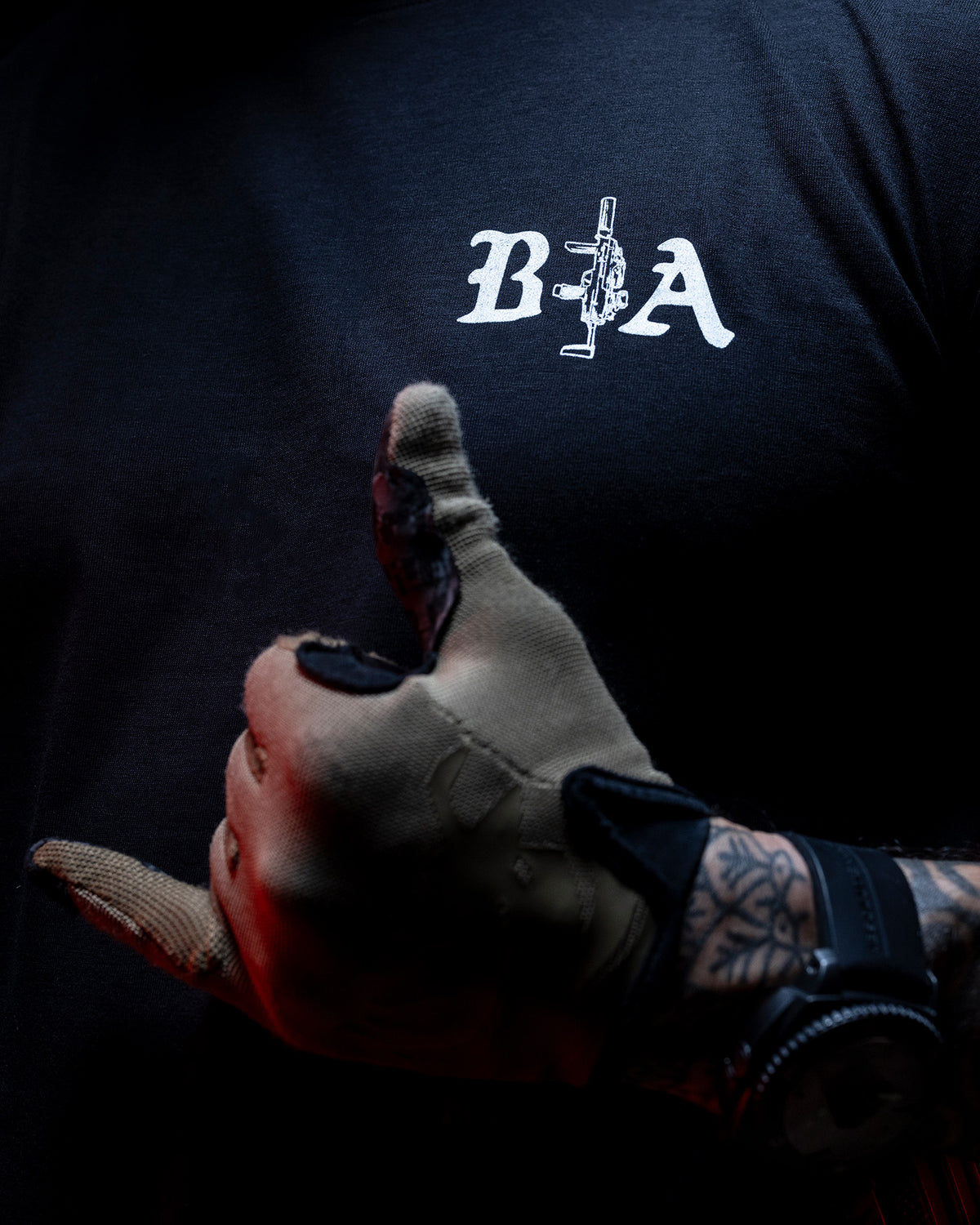 Gloved hand reaching forward, the B|A chest logo on the black Hard Times T-Shirt behind it