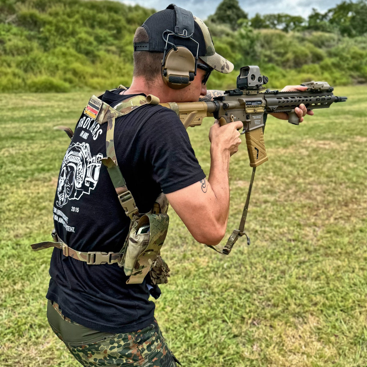 Man in a black Brothers in Arms T-Shirt firing a rifle with a plate carrier outdoors