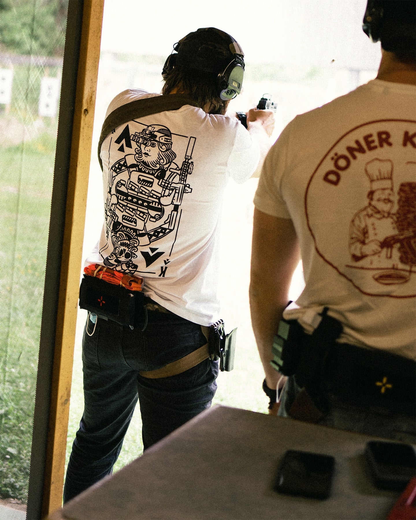 Two people at a shooting range, one in the white King of Spades T-Shirt, one in a Döner Kalash shirt #color_white #in_field