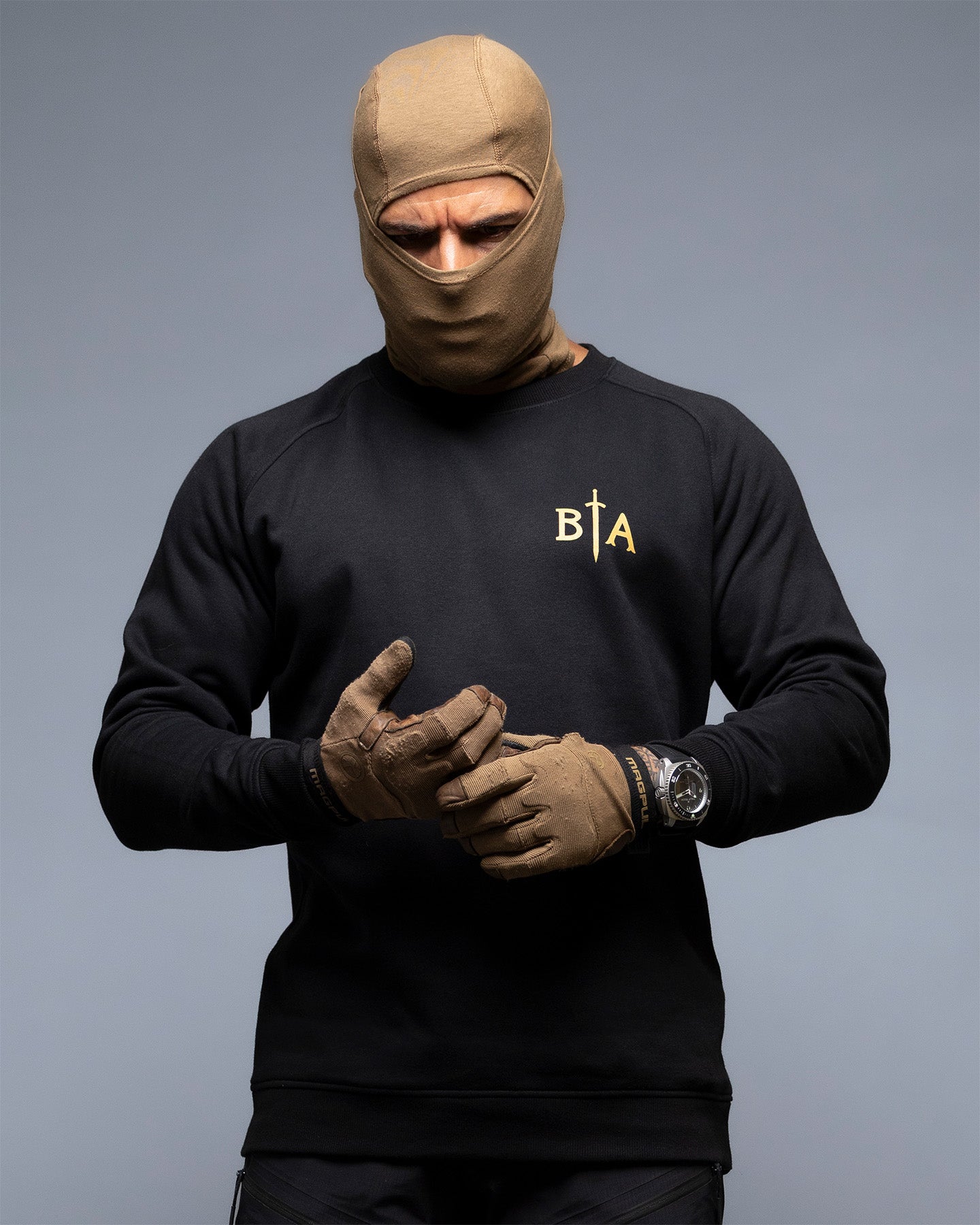 Person wearing a black sweatshirt with a logo, brown gloves, and a beige balaclava on a gray background. #color_black