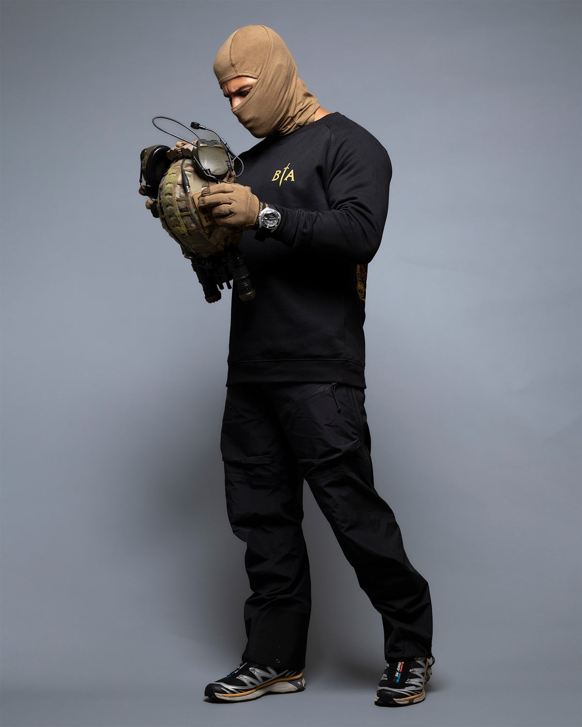 Man in a balaclava holding a helmet at full length, wearing the black No Fear Crewneck