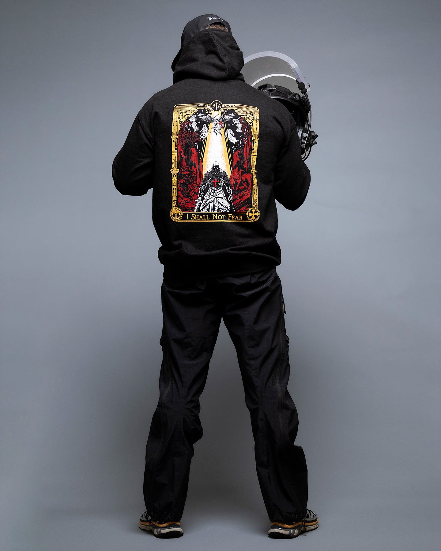 Person wearing a black hoodie with a graphic design on the back against a gray background. #color_black