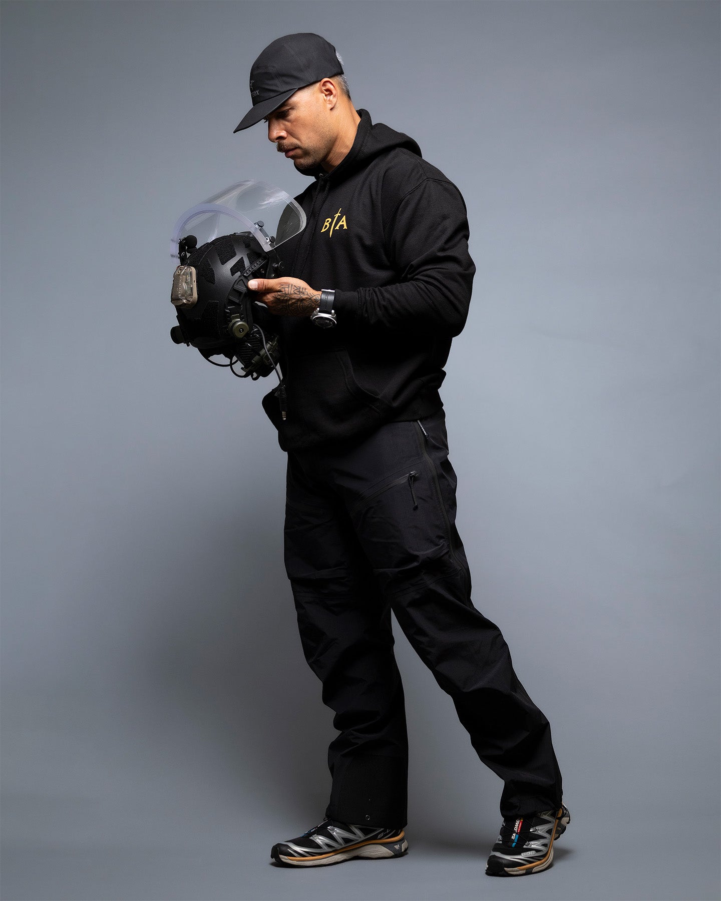 Person in black tactical gear holding a helmet against a gray background. #color_black