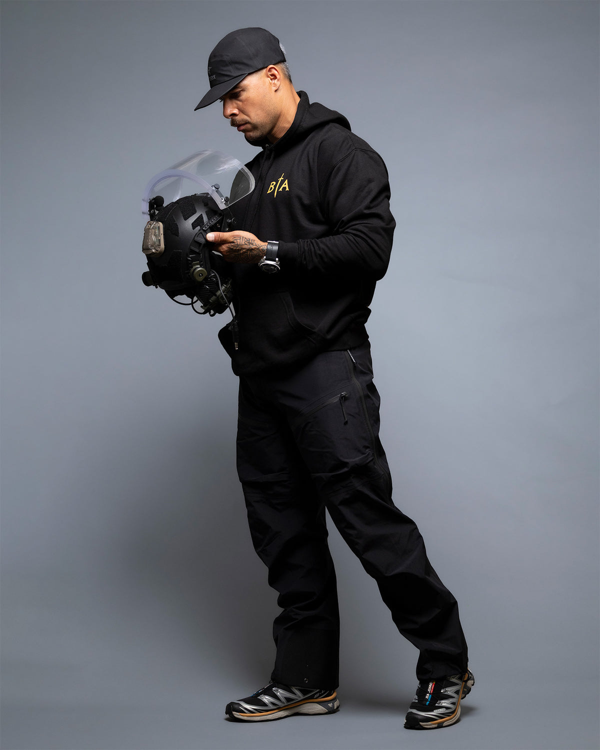 Man in a cap at full length holding a visor helmet, wearing the black No Fear Hoodie