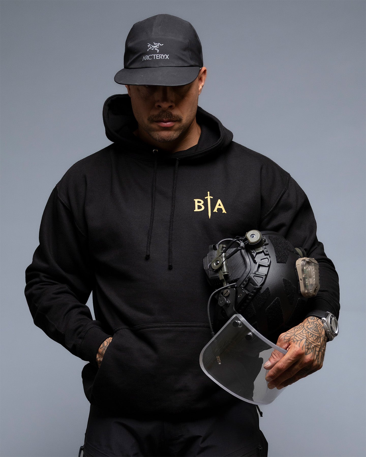Person wearing a black hoodie with a logo, holding a helmet against a gray background. #color_black