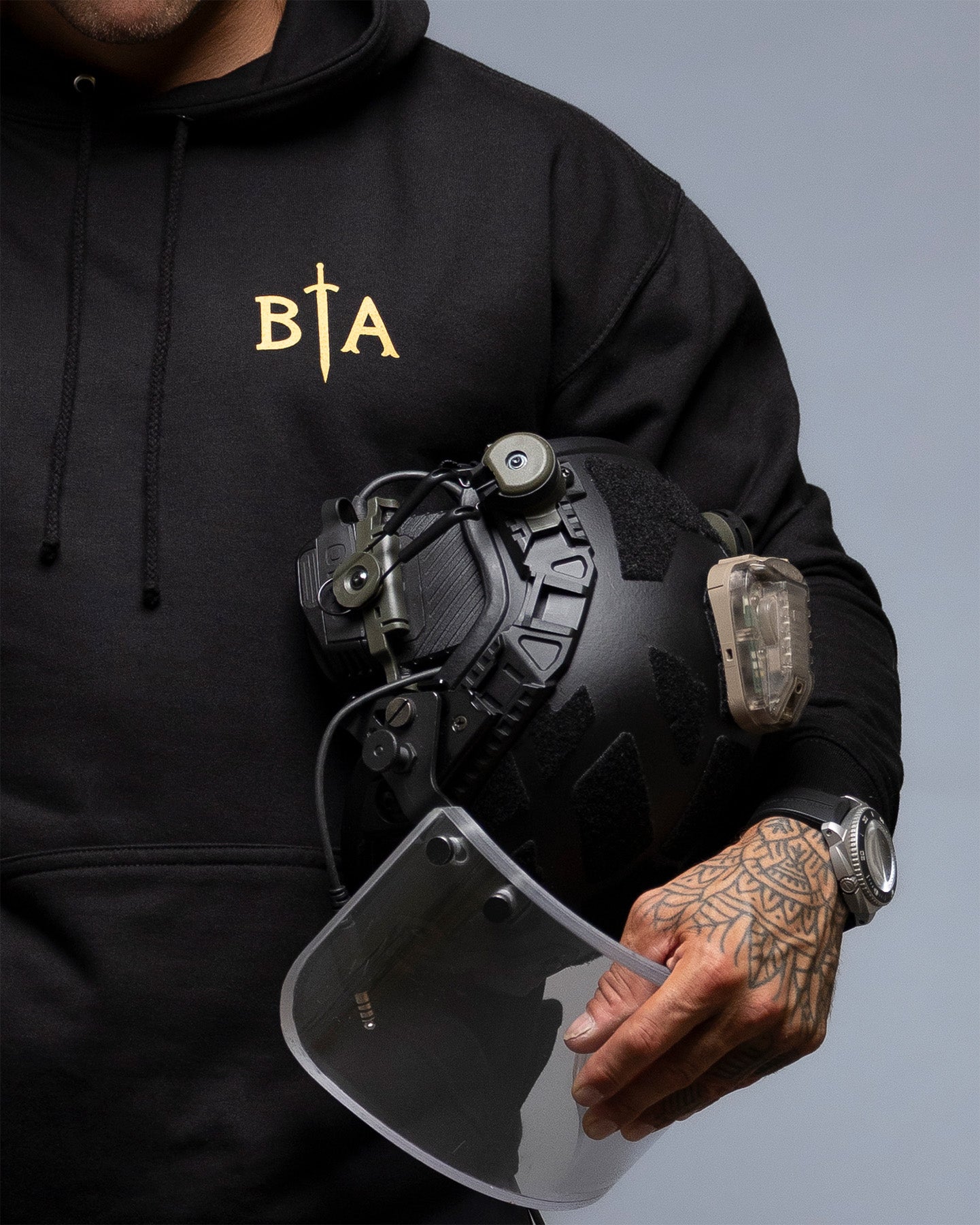Person wearing a black hoodie with 'BIA' logo, holding a tactical helmet and face shield. #color_black