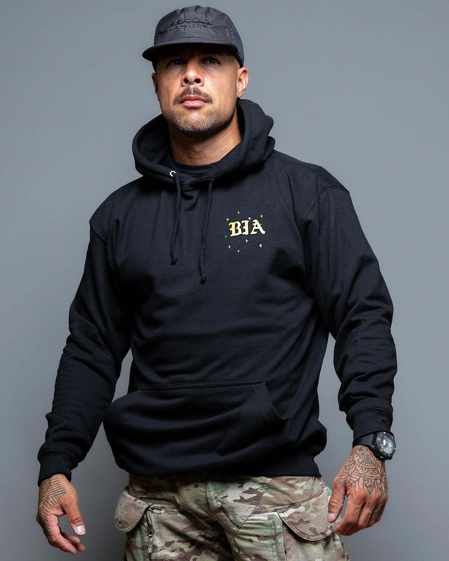 Man wearing a black hoodie with 'BIA' logo and camouflage pants on a gray background. #color_black
