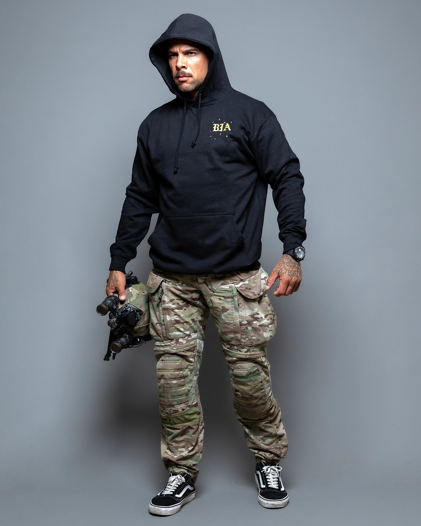 Person wearing a black hoodie with a logo and camouflage pants on a gray background. #color_black