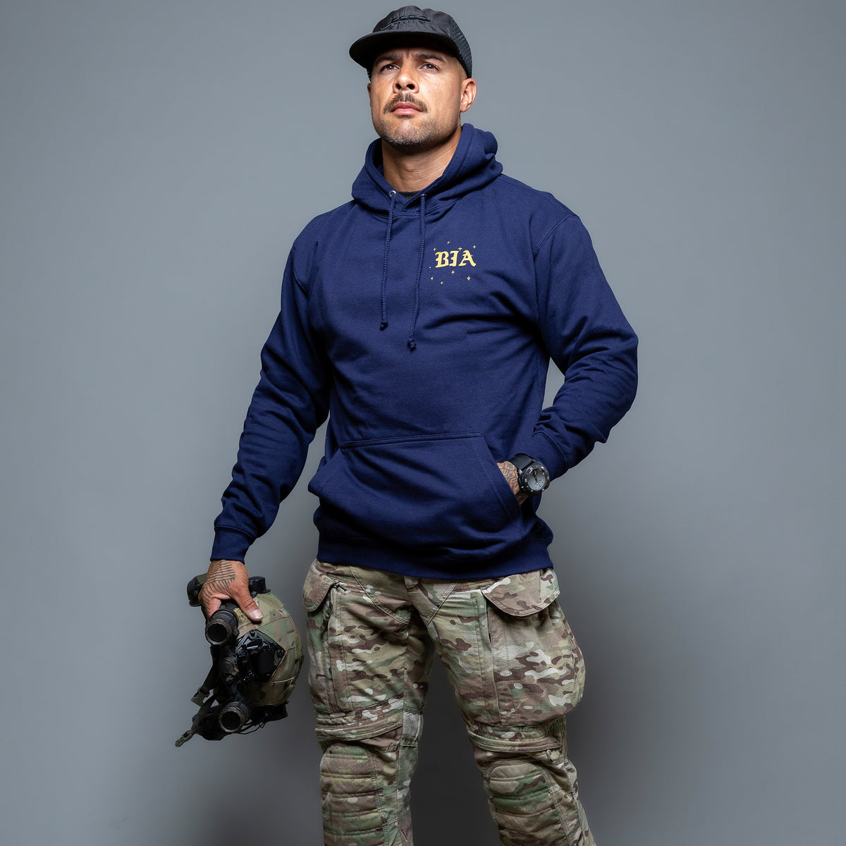 Man in a cap holding a helmet, wearing the navy Eyes On The Stars Hoodie with the gold chest print