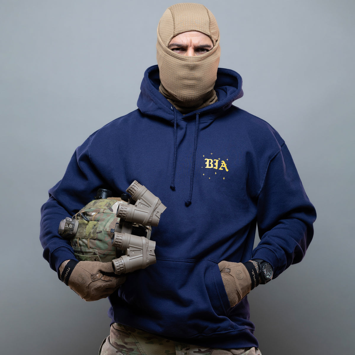 Man in a balaclava holding a helmet with night vision, wearing the navy Eyes On The Stars Hoodie