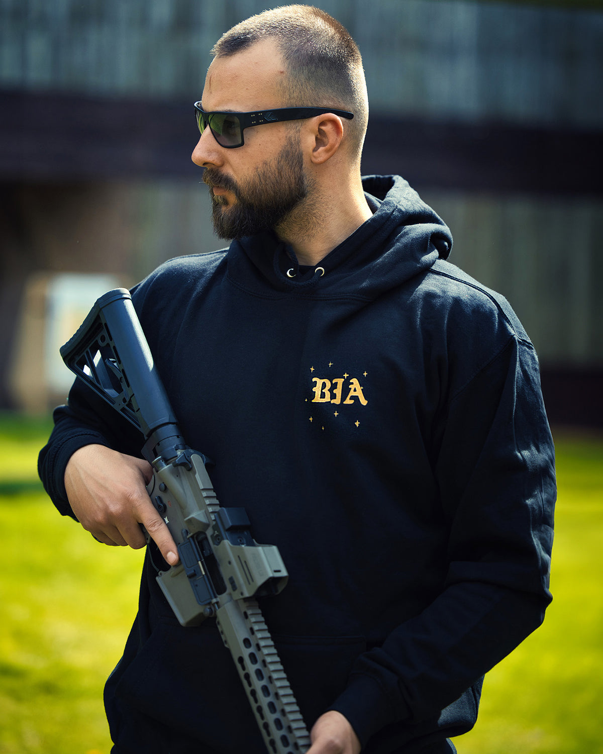 Man in sunglasses holding a rifle outdoors, wearing the black Eyes On The Stars Hoodie with the gold chest print