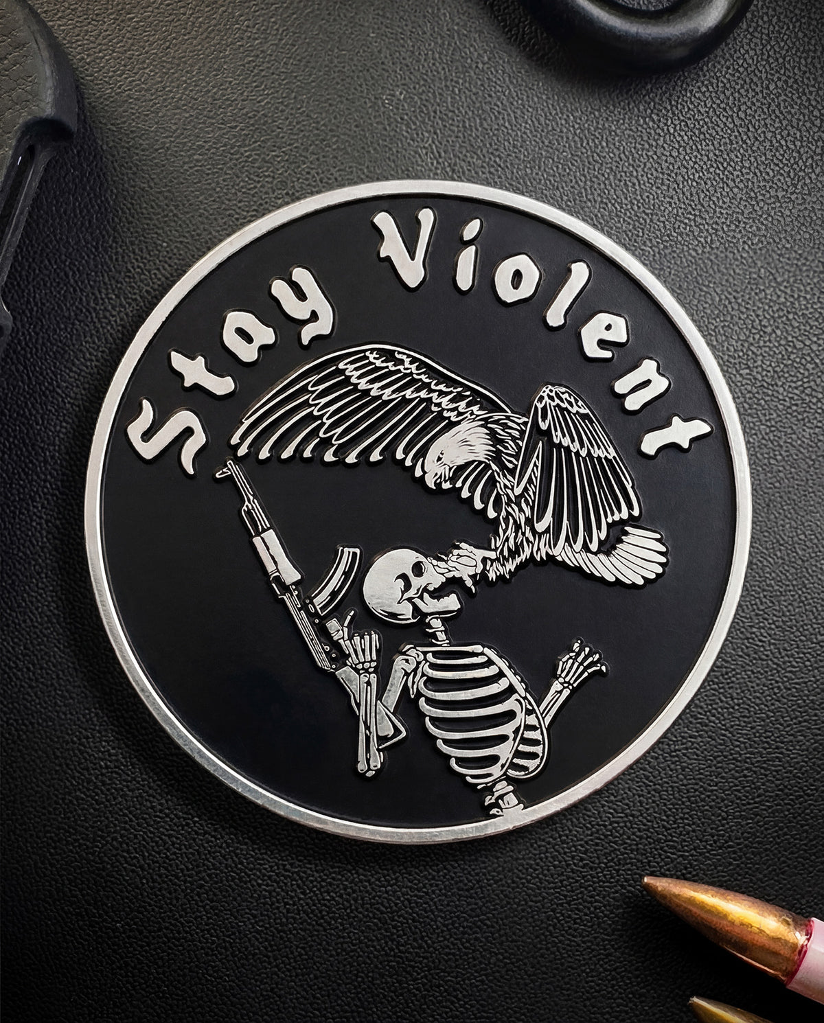 Close-up of the Stay Violent coin front with the winged skeleton, cartridge beside it