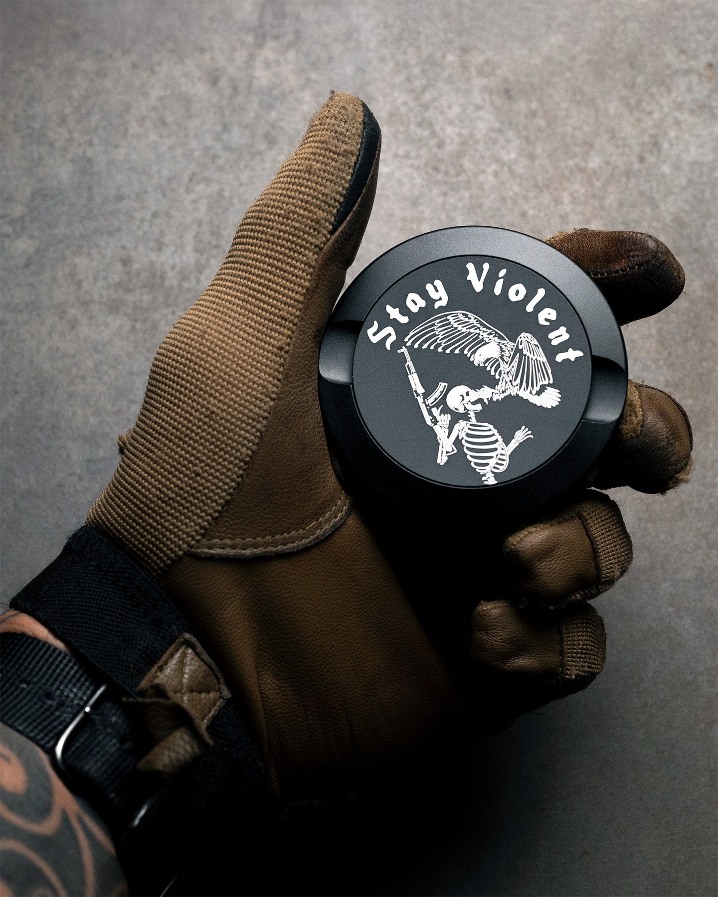 Hand wearing a brown glove holding a black pouch can with 'Stay Violent' text and skeleton design on a gray background. #in_field